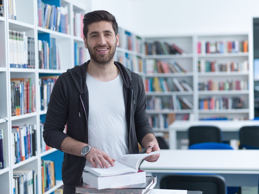 A smiling man in a library holding a question bank book for MCCQE Part 1.