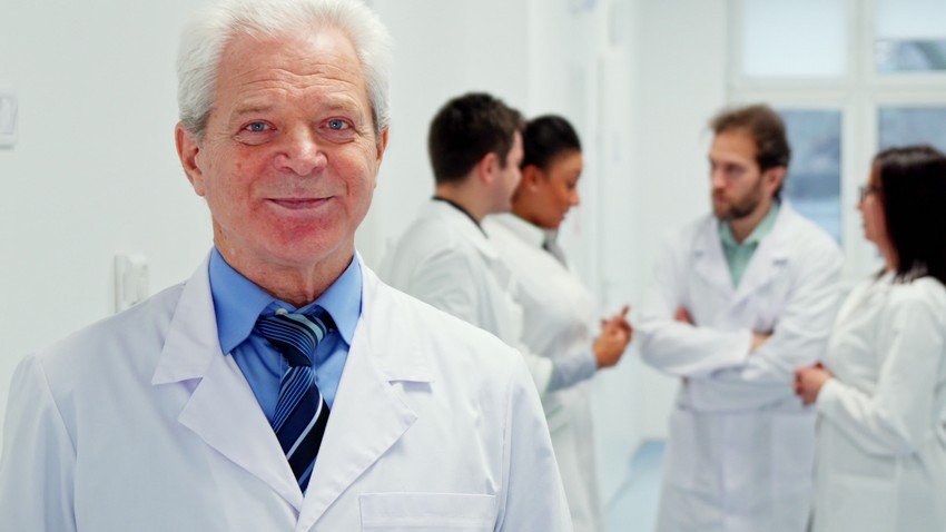 Senior doctor in white coat with colleagues discussing PLAB 1 in the background.