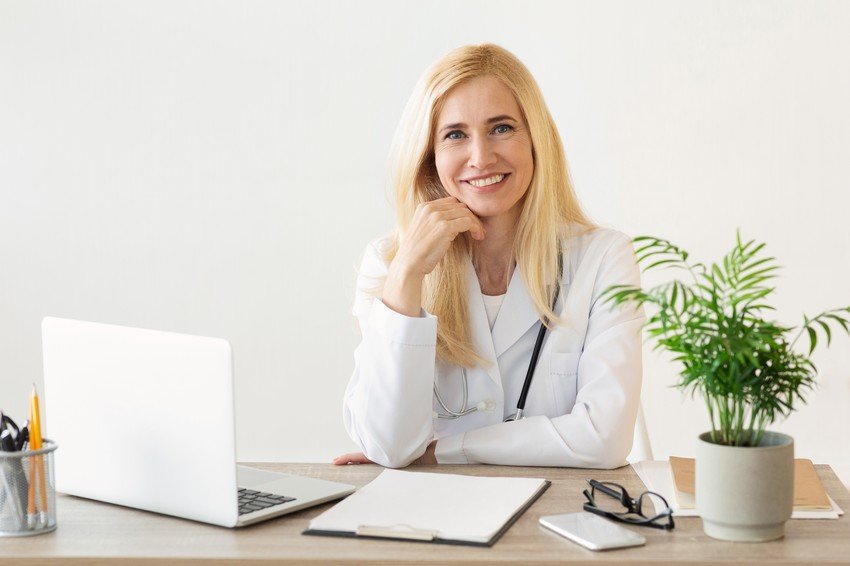 Doctor smiling at her desk with laptop and office supplies, preparing for PLAB.
