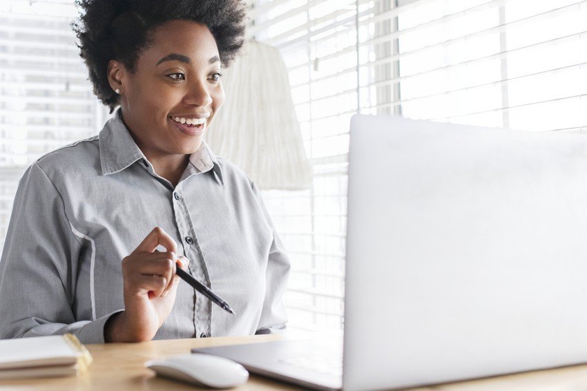 A smiling woman working on a PLAB question bank on her laptop by a window with blinds.