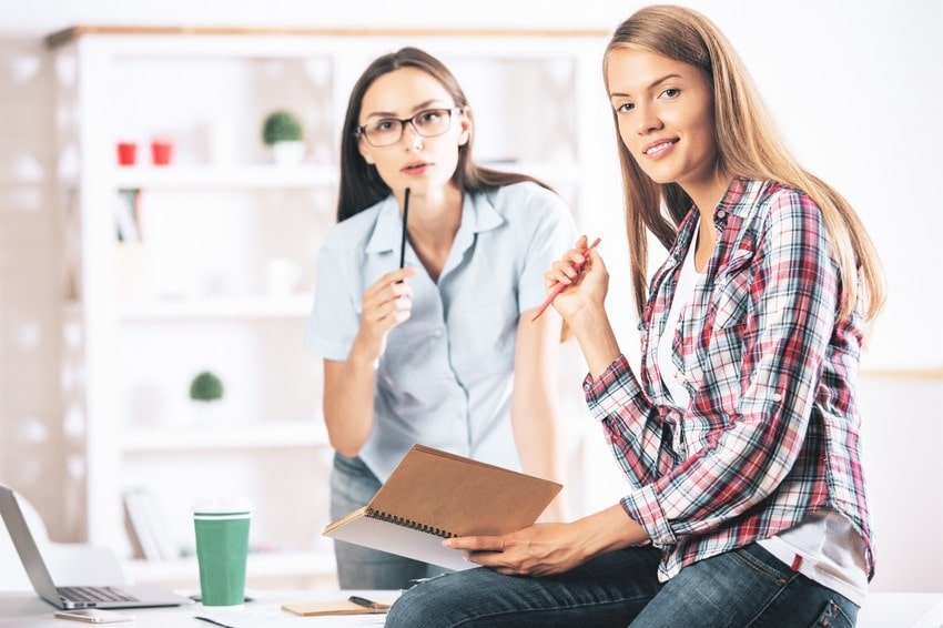Two medical students in a bright office, one wearing glasses and holding a notebook, the other sitting on a desk with a laptop and a coffee cup nearby, both appear engaged in a discussion about their MCCQE1.