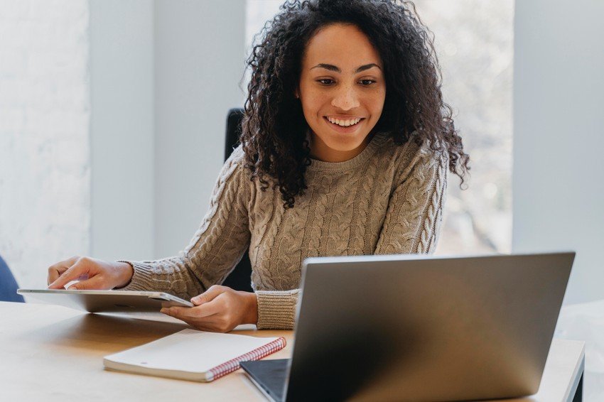 A smiling medical student with curly hair using a tablet and laptop at a desk for MCCQE1 exam preparation, with a notebook beside her.