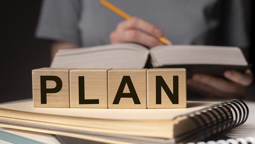 Person writing in a notebook next to wooden blocks spelling out "plan" on top of medical books.