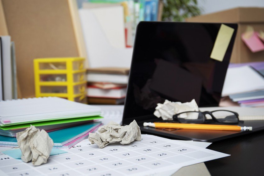 A cluttered office desk with an open laptop, glasses, crumpled paper, and a calendar among scattered documents and a packed moving box for MCCQE1 exam preparation.