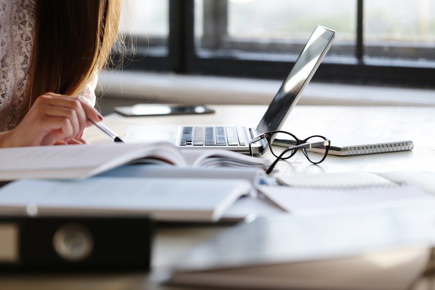 Woman studying for the MCCQE1 exam with books and laptop at a desk, writing notes, with glasses lying near her computer.