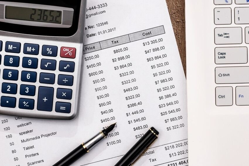 A calculator, a pen, and a paper with columns of price, tax, and cost amounts are on a wooden desk next to a laptop keyboard, illustrating how such tools are essential in understanding the role of MCCQE1 in doctor salaries.
