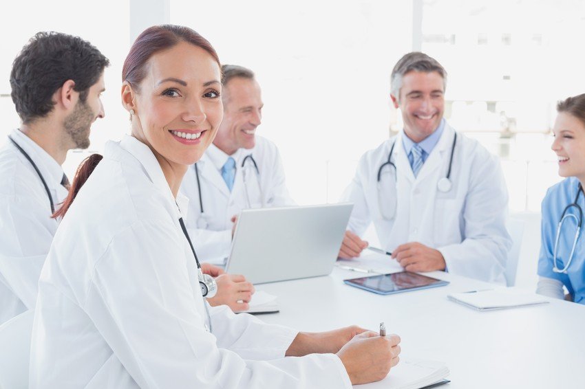 A group of medical professionals, including female doctors, smiling and sitting around a table with laptops in meetings, saying women need to break the glass ceiling with MCCQE1.