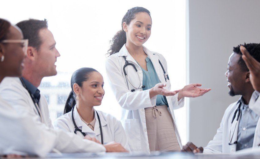 A group of doctors in lab coats, some with stethoscopes, sit at a table engaged in a discussion about doctor salary. One standing doctor gestures while speaking to the group about MCCQE1.