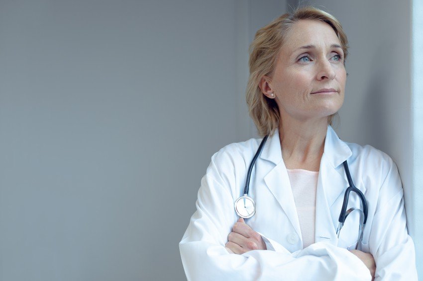 A middle-aged female doctor in a white coat with a stethoscope, looking thoughtful and gazing upwards, thinking about encouraging women to break the glass ceiling with MCCQE1.