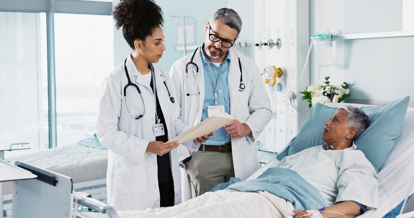 Two doctors, possibly discussing doctor salaries, stand by a hospital bed, deliberating over a medical chart with a patient lying in bed. Medical equipment is visible in the background, hinting at the rigorous training from their medical residencies.