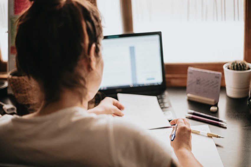 A person sits at a desk working on a laptop with papers and a pen in hand, preparing to overcome MCCQE1 exam anxiety using Ace QBank. Various office supplies are scattered around, including a calendar and a small cactus.