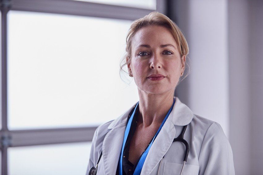 A confident female doctor with a stethoscope around her neck is standing in front of a window in a medical office, thinking about Ace QBank.