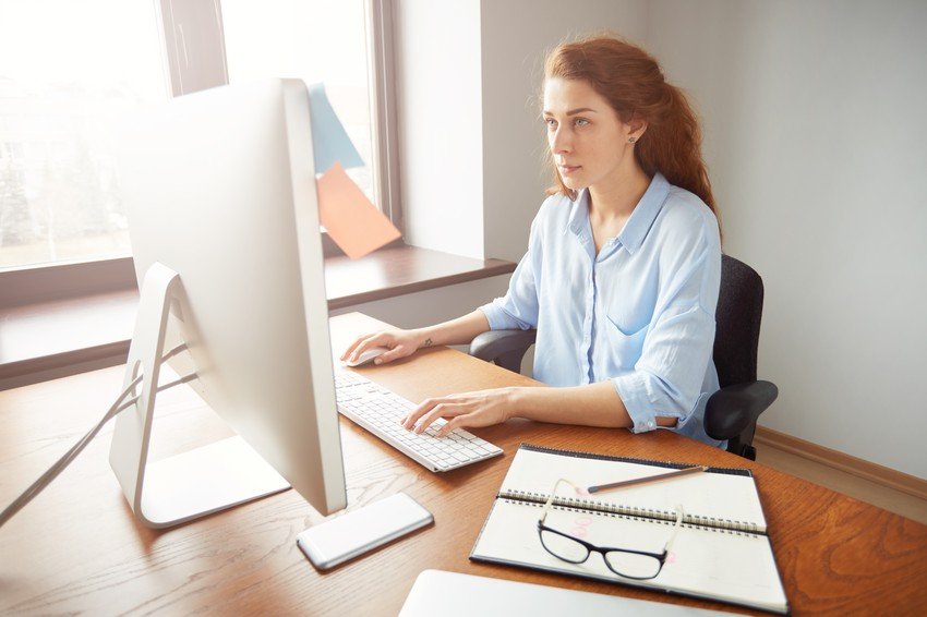 A woman is working on a computer at a desk with a notebook, pen, and glasses. She is sitting in a modern office with large windows, likely preparing to tackle her MCCQE1 exam anxiety with resources like Ace QBank.