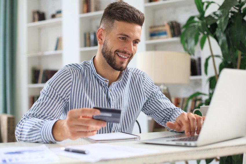 A man in a striped shirt uses a laptop while holding a credit card at a desk, possibly setting up his Ace QBank demo account. Bookshelves and a potted plant are visible in the background.