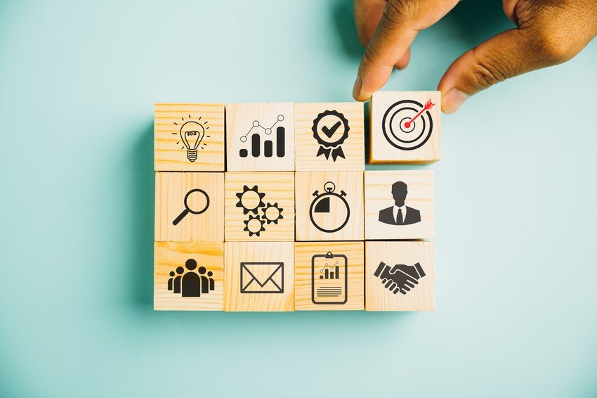 A hand arranges blocks featuring icons for ideas, analytics, achievement, target, search, settings, teamwork, MCCQE1 preparation with Ace QBank, email, task list, and handshake on a light blue background.