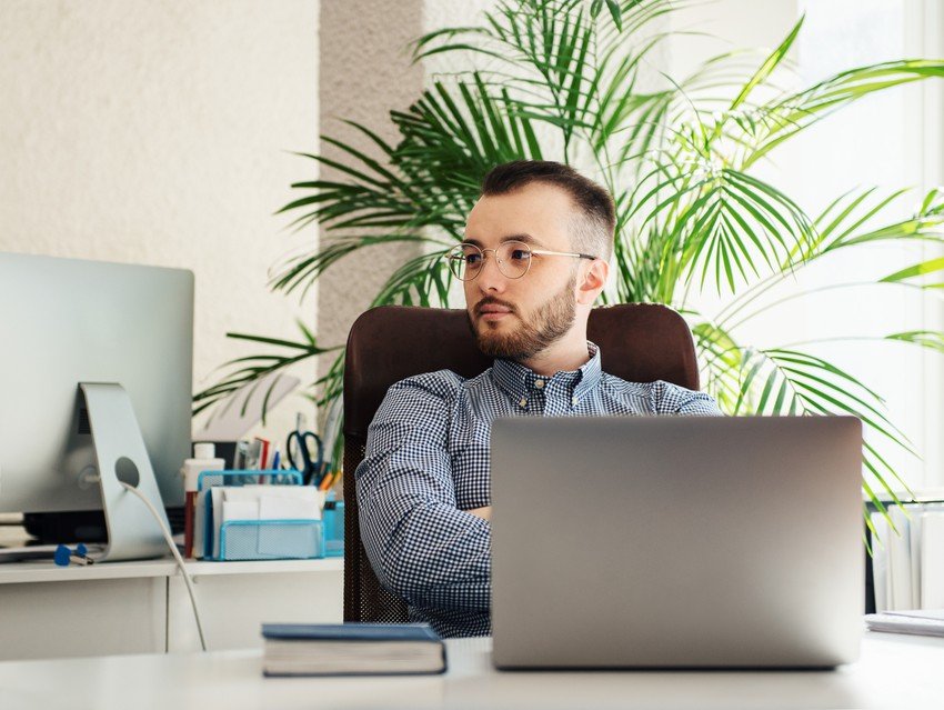 A man with glasses sits at a desk with a laptop in front of him, glancing to the side. Amidst a background of office supplies and a large plant, he seems deep in thought, possibly dealing with MCCQE1 exam anxiety. Maybe he's considering using Ace QBank for his MCCQE1 prep.