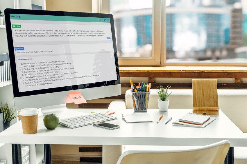 A computer on a white desk displays a web page on how to practice questions for the MCCQE Part I. Surrounding items include a green apple, a notepad, a pen, potted plants, and a clipboard. A window in the background provides natural light.