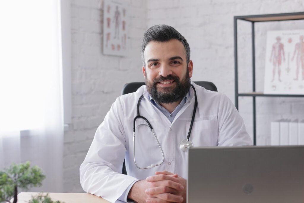 A bearded man in a white coat with a stethoscope around his neck sits at a desk and smiles at the camera. Medical charts, MCC objectives, and Ace QBank materials are visible on the wall in the background.