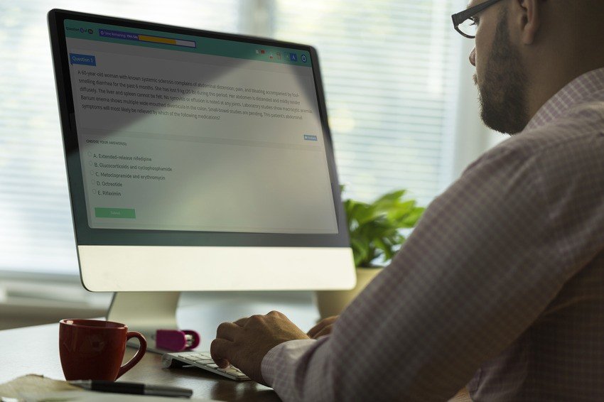A person wearing glasses and a plaid shirt is working on a computer, focused on MCCQE1 exam preparation using Ace QBank. The monitor displays a webpage with text and multiple-choice questions. On the desk, there’s a red mug, a small flashlight, and a pen.