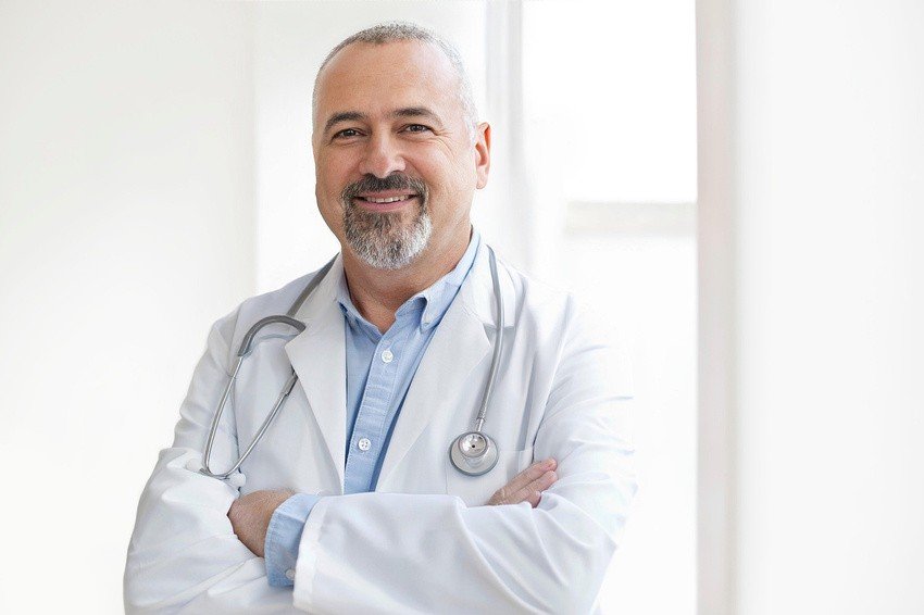 A middle-aged man in a white coat with a stethoscope draped over his shoulders stands smiling with arms crossed against a light background, exuding confidence as if he's mastered Ace QBank for MCCQE1 exam preparation.