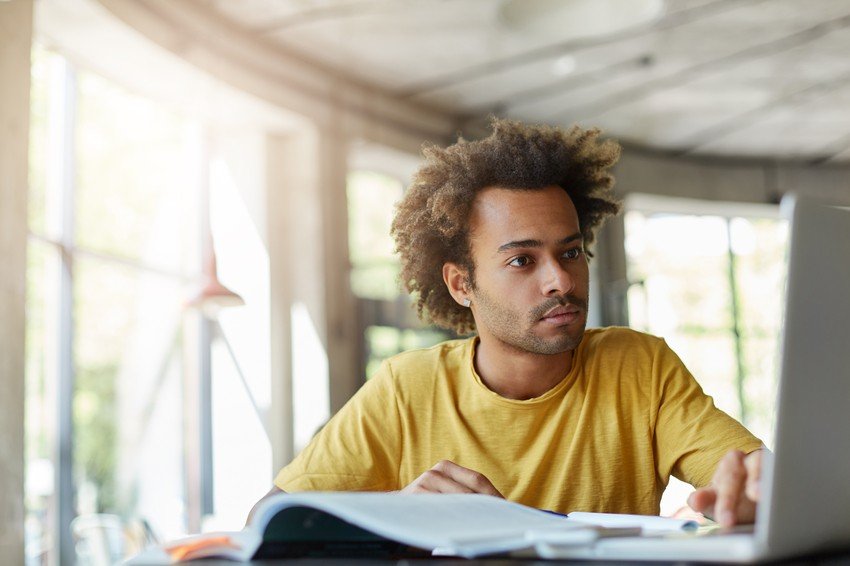 A person with curly hair in a yellow shirt is sitting at a desk, focusing on a laptop screen displaying Ace QBank for MCCQE1 exam preparation, with open books and notebooks nearby in a bright room.