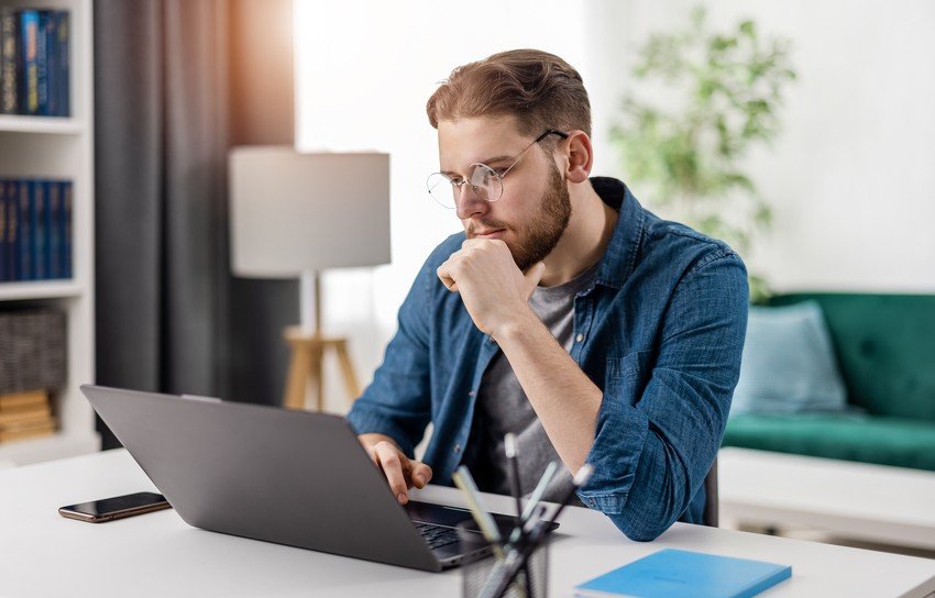 A man with glasses and a beard sits at a desk, focused on his laptop researching how to practice questions for the MCCQE Part I. He is wearing a blue denim shirt and resting his chin on his hand. A smartphone, an Ace Qbank guide, and a pen holder are on the desk.
