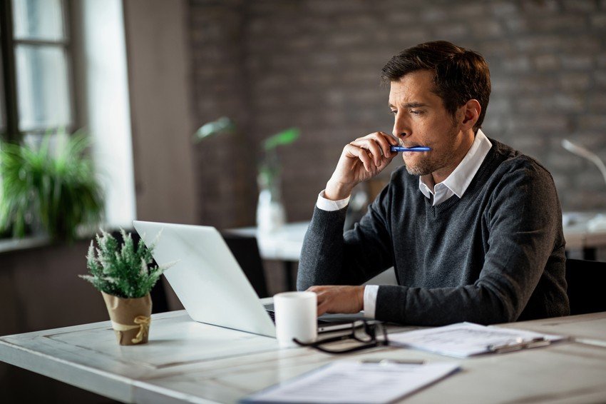 A man sits at a desk, looking pensively at a laptop screen, with "Ace Qbank" displayed. Holding a pen to his lips, he ponders how to practice questions for the MCCQE Part I. A potted plant, coffee mug, and paperwork are scattered across the desk.