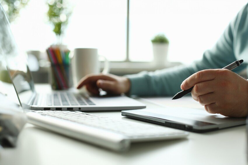 Person using a laptop and holding a pen, with a smartphone, keyboard, and office supplies on the desk in a bright workspace. They're preparing for the MCCQE Part I by using an Ace QBank online question bank to practice questions effectively.