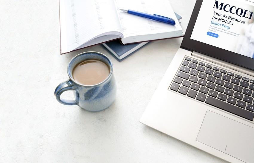 A laptop displaying the Ace QBank Website for the MCCQE1 exam prep on a table next to a notebook, a blue pen, and a blue ceramic mug filled with coffee.
