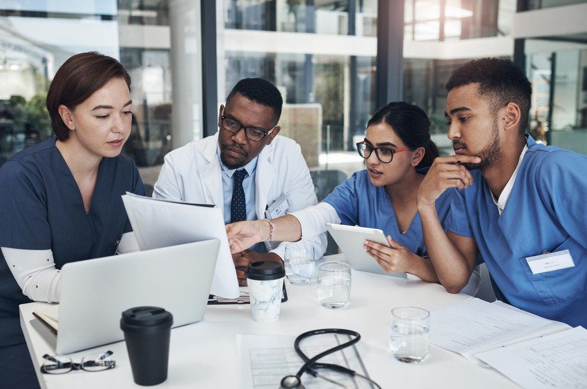 Four medical professionals in scrubs and lab coats discuss MCCQE1 documents around a table with laptops, a stethoscope, and drinks in a modern office setting. The presence of Ace QBank resources aids their comprehensive review and collaborative approach to medical excellence.
