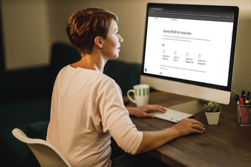 A woman sits at her desk, gazing intently at a computer displaying session dates for MCCQE1. Beside her, a coffee mug and a small plant add comfort as she prepares with Ace QBank.