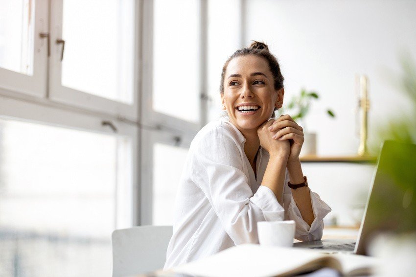 A woman in a white shirt is seated at a desk near a window, smiling confidently as she prepares to crush 2025 MCCQE1 on her first try. A laptop and a small cup beside her reflect dedication and focus, tools of success with Ace QBank.