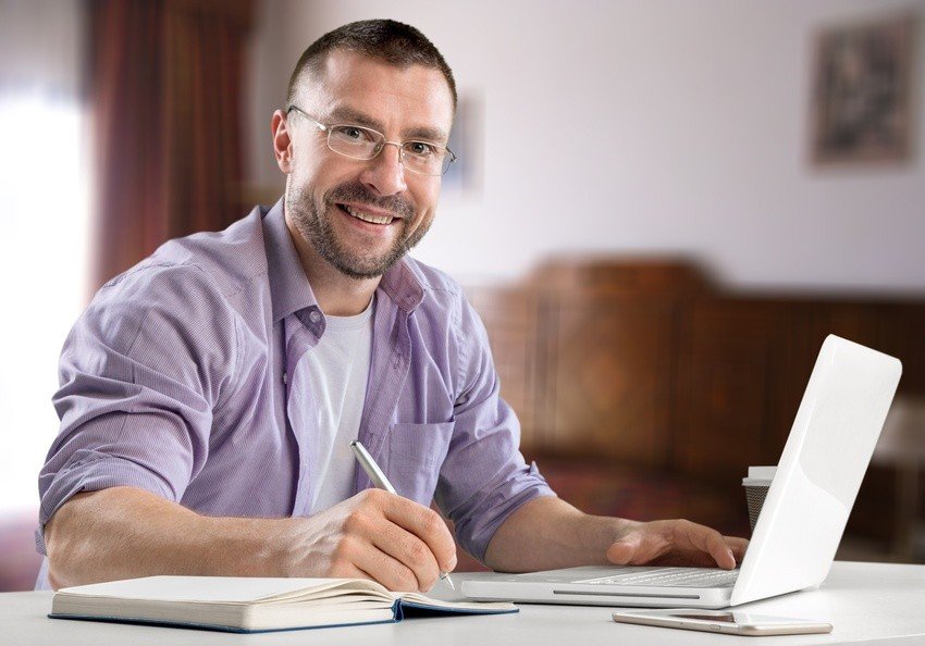 A man wearing glasses smiles as he sits at his desk with a laptop, notebook, and pen, ready to ace the MCCQE1 with Ace QBank and crush the 2025 exam on his first try.