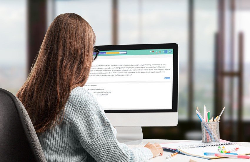 A woman with long brown hair sits at a desk practice questions on her computer with Ace QBank, perhaps after researching AMC exam dates. She wears a light blue sweater.