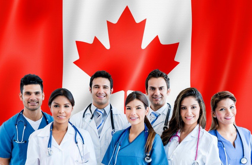 A diverse group of healthcare professionals, confident and smiling in their medical attire, stand proudly in front of a Canadian flag. They're ready to utilize Ace QBank for their 2025 MCCQE Part 1 prep & resources, embracing the MCC objectives with dedication.