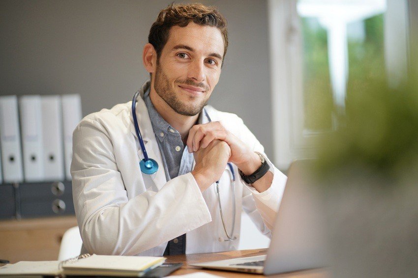 A doctor in a white coat with a stethoscope around his neck sits at a desk, smiling confidently as he reviews AMC exam preparation materials on his laptop, poised to master the upcoming AMC exam dates with Ace QBank resources by his side