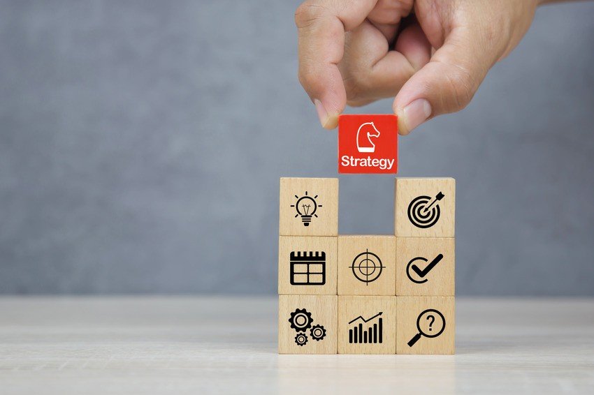 A hand places a red block labeled "AMC exam strategy" atop wooden blocks adorned with business icons like lightbulb, target, calendar, cogwheels, graph, and question mark—an essential move in managing AMC exam dates with precision.