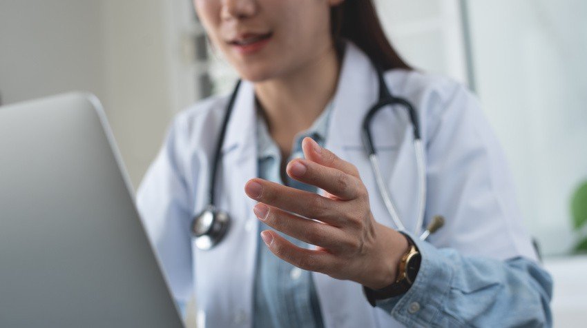 A doctor in a white coat, with a stethoscope around their neck, gestures thoughtfully while reviewing MCCQE1 sample questions on a laptop.
