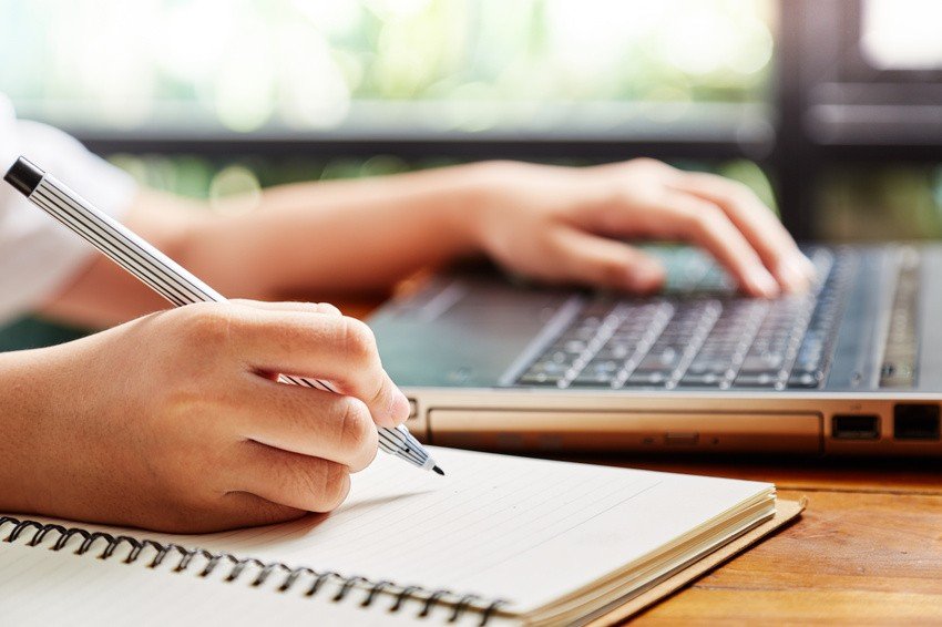 A person is diligently jotting down notes with a pen in their notebook, using a laptop placed on a wooden table by the window, where blurred greenery peeks through. They are likely preparing for MCCQE Part 1 with sample questions spread across their screen.