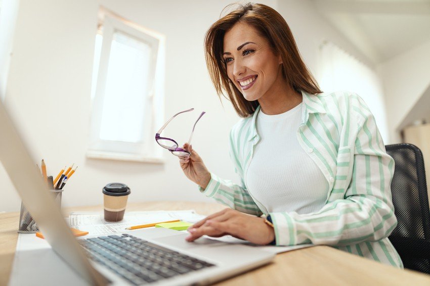 A woman smiles while using her laptop at a desk, holding glasses in one hand, clearly engaged in her 2025 MCCQE part 1 prep. A takeaway coffee cup and stationery lie nearby, perhaps with Ace QBank aiding her MCCQE1 journey.