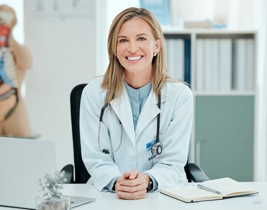 A female doctor in a white coat sits at a desk, smiling, with a stethoscope around her neck and an open notebook in front of her, ready to tackle MCCQE1 prep and review MCC objectives using Ace QBank.