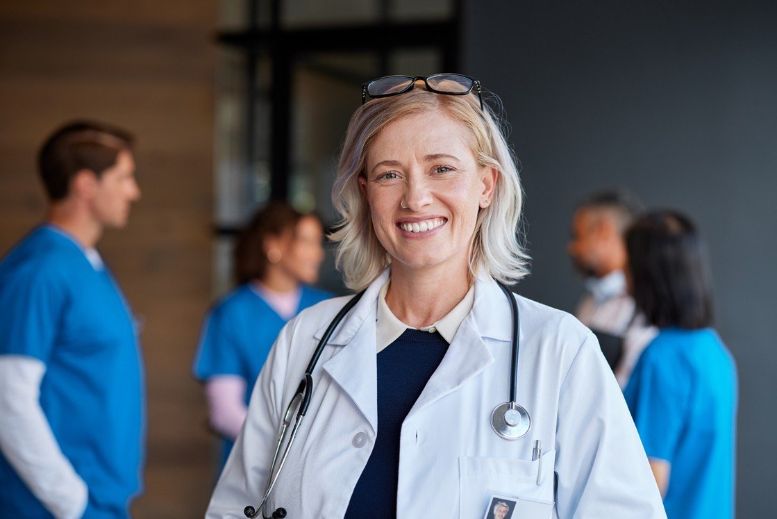 A smiling doctor with a stethoscope stands in the foreground, while a group of medical professionals in blue scrubs discuss MCCQE1 exam preparation in the background.