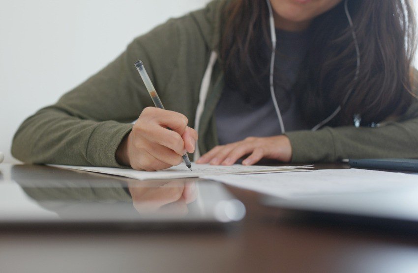 A person in a green hoodie is writing on paper at a desk, earphones in and a tablet nearby. They're fully immersed in their MCCQE1 Prep, using a Qbank to ensure comprehensive understanding for the MCCQE Part 1 exam.