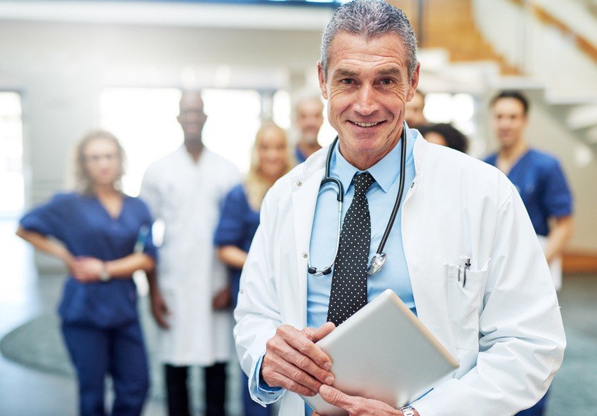 A doctor in a white coat, perhaps pondering strategies for MCCQE1 Prep, holds a folder while standing before a group of medical professionals in scrubs and lab coats.