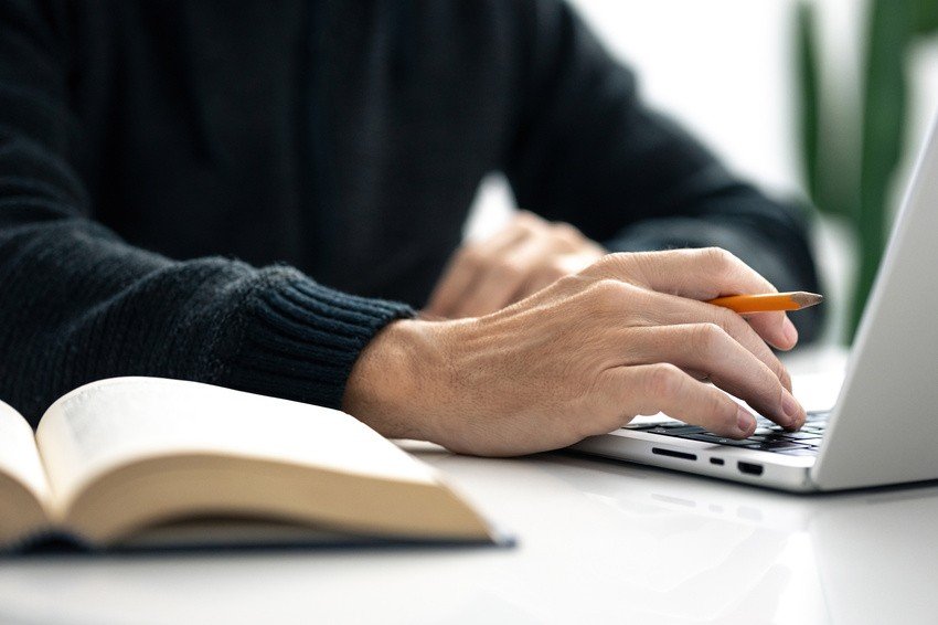 A person is engrossed in typing on a laptop with an open book nearby on a white desk, diving into MCCQE1 Prep using the Qbank for improved study efficiency.