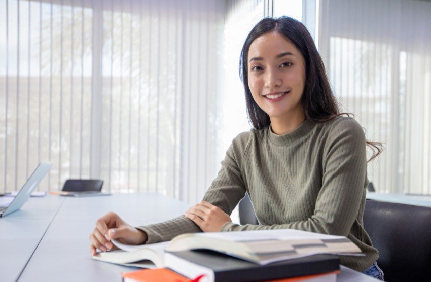 A woman sits at a table with books, wearing a long-sleeve green top, likely immersed in her MCCQE1 Prep using Ace QBank to boost her confidence and readiness.
