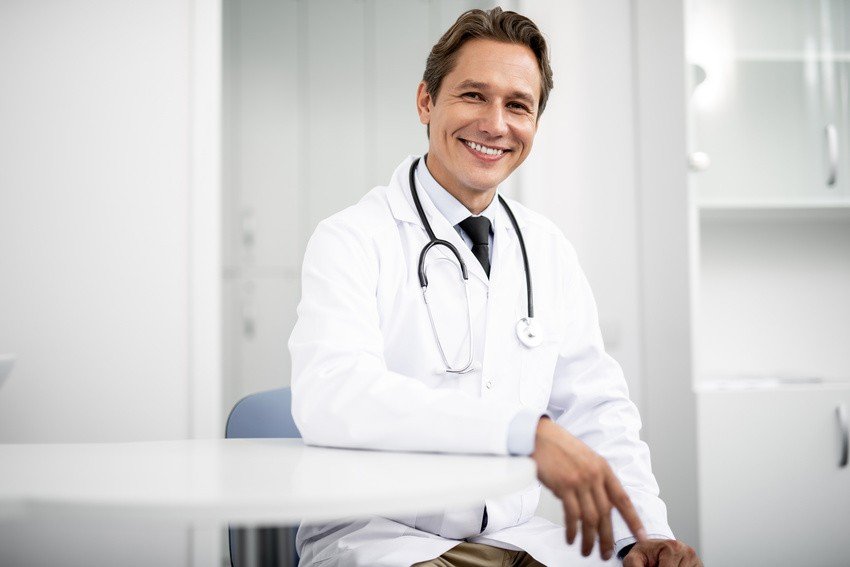 A male doctor in a white coat with a stethoscope around his neck sits and smiles in a bright, modern medical office, confident after using Ace QBank for MCCQE1 Exam Prep.