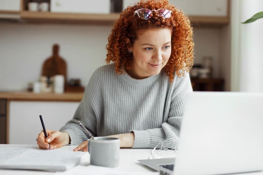 A woman with curly red hair sits at a table, writing in a notebook and looking at a laptop—focused on her MCCQE Part 1 Prep. A gray mug and some earphones are next to her as she studies for the MCCQE1 exam.