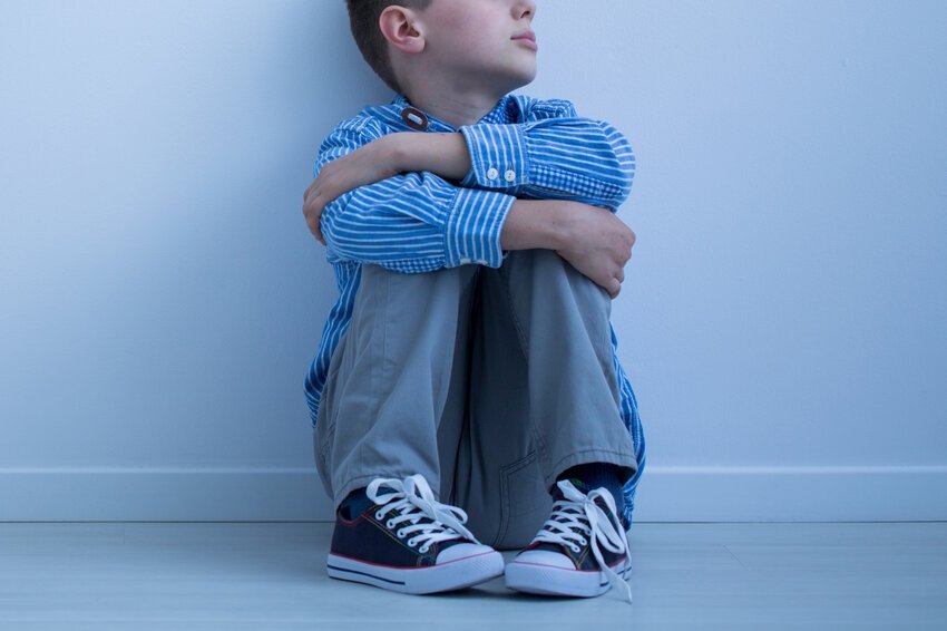 A young boy in a blue plaid shirt and gray pants sits on the floor with his arms around his knees, reflecting common behaviours seen in children with autism spectrum disorder. Ace QBank – Clinical edge