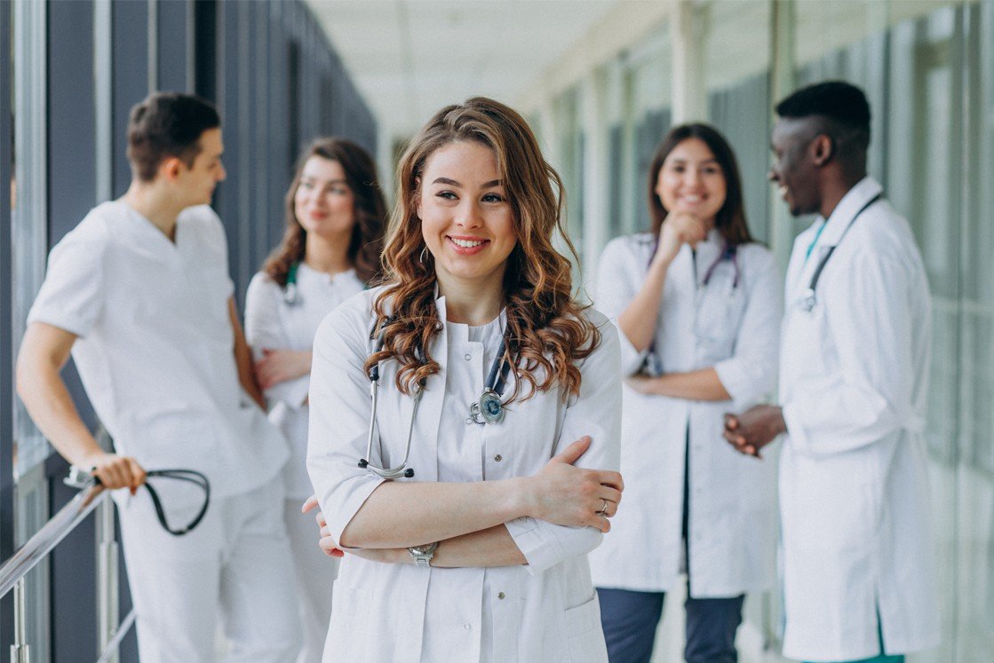 A group of five medical professionals in white coats and scrubs stand in a hallway, with one woman in front smiling at the camera—ready for their MCCQE1 exam after thorough Ace QBank and MCCQE Part 1 Prep.